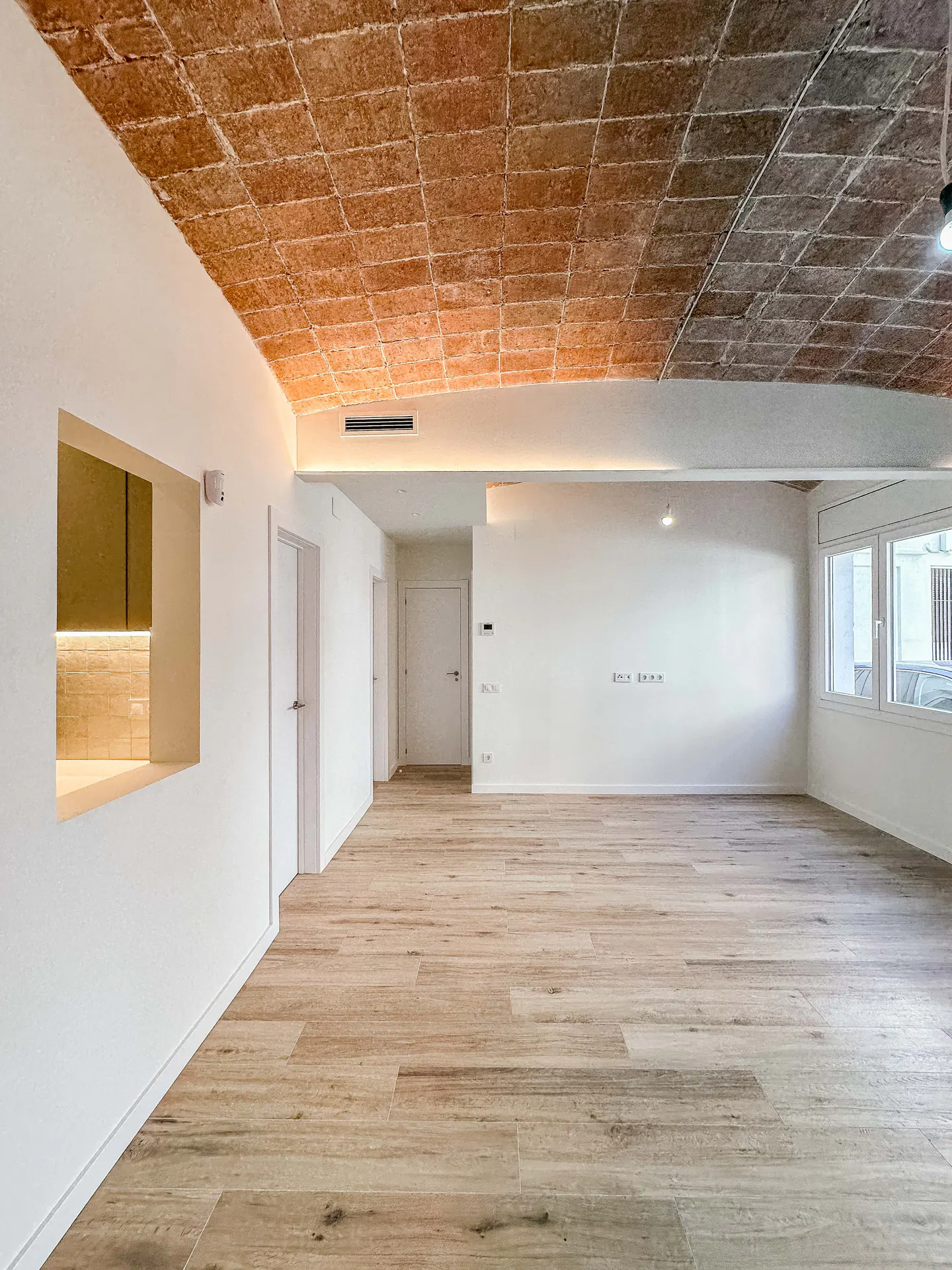 Bright, empty room with light wood floors, white walls, and a brick-patterned ceiling. Windows on the right let in natural light.