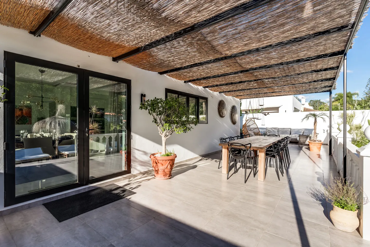 Outdoor patio with a dining table and chairs under a thatched roof. Black framed glass doors lead inside. Potted plants add greenery.