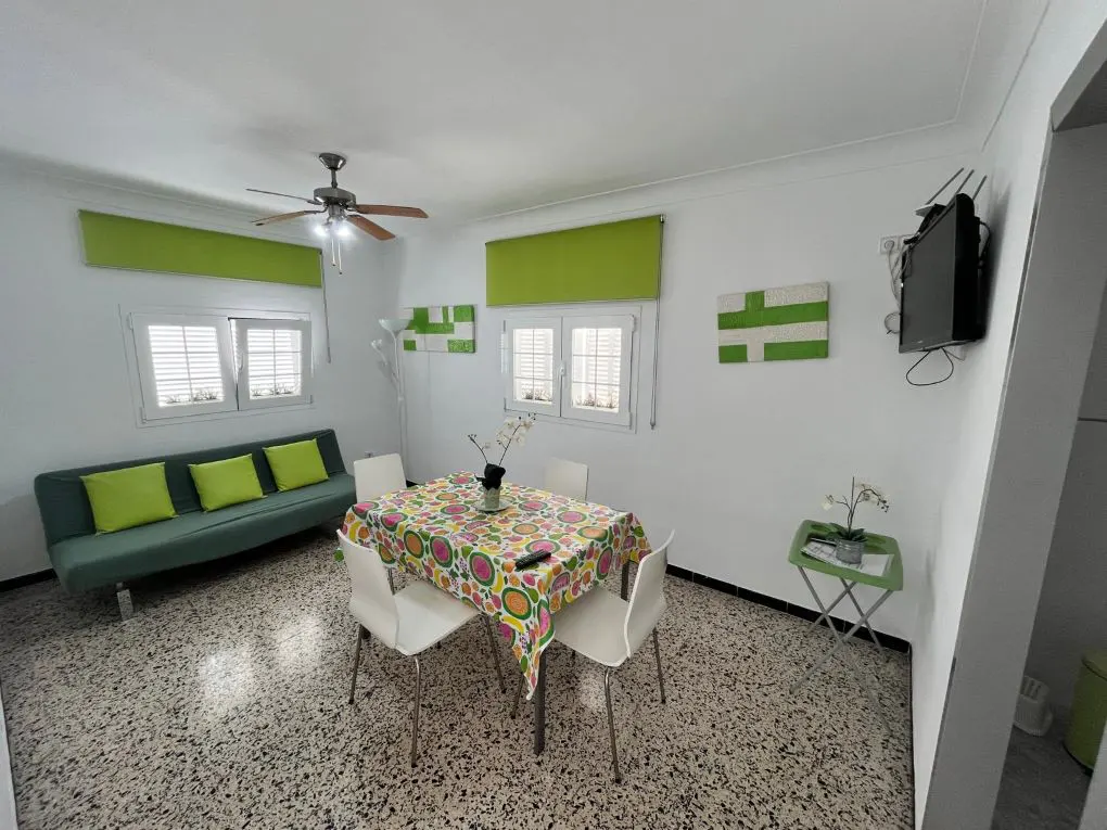 Bright living room with white walls, terrazzo floor, green sofa, and a dining table with a colorful tablecloth. A TV hangs on the wall.