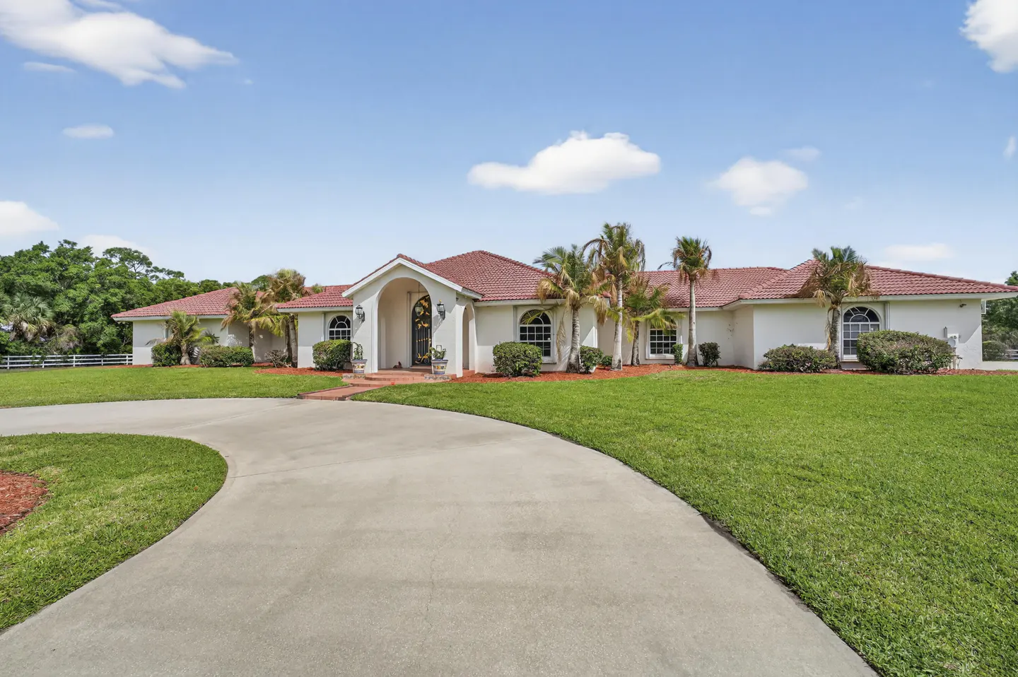 A single-story white house with a red tile roof, palm trees, and a curved driveway on a sunny day.