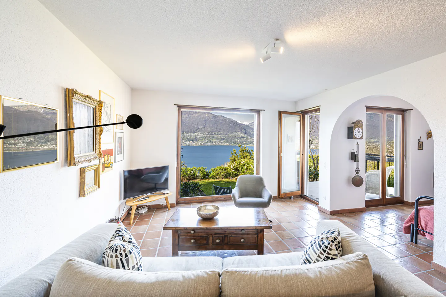 Bright living room with white walls, terracotta tile floor, and a view of a lake and mountains through a large window. A beige sofa faces a dark wood coffee table.