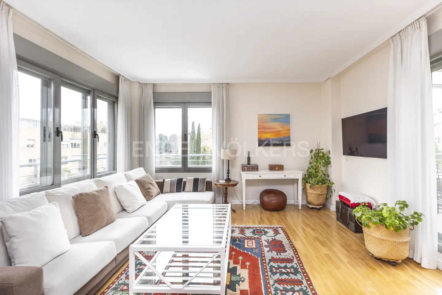 Bright living room with a white sofa, glass coffee table, and colorful rug on a wood floor. Large windows with sheer curtains.