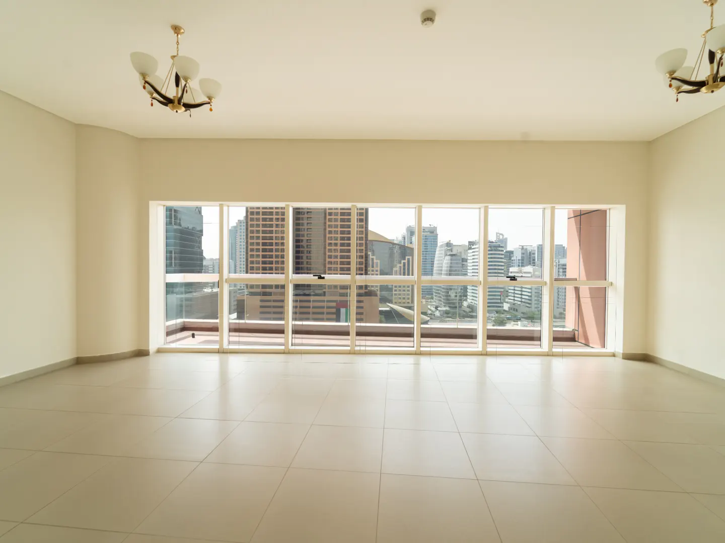 Empty room with beige walls, tiled floors, two chandeliers, and large windows overlooking a city skyline.