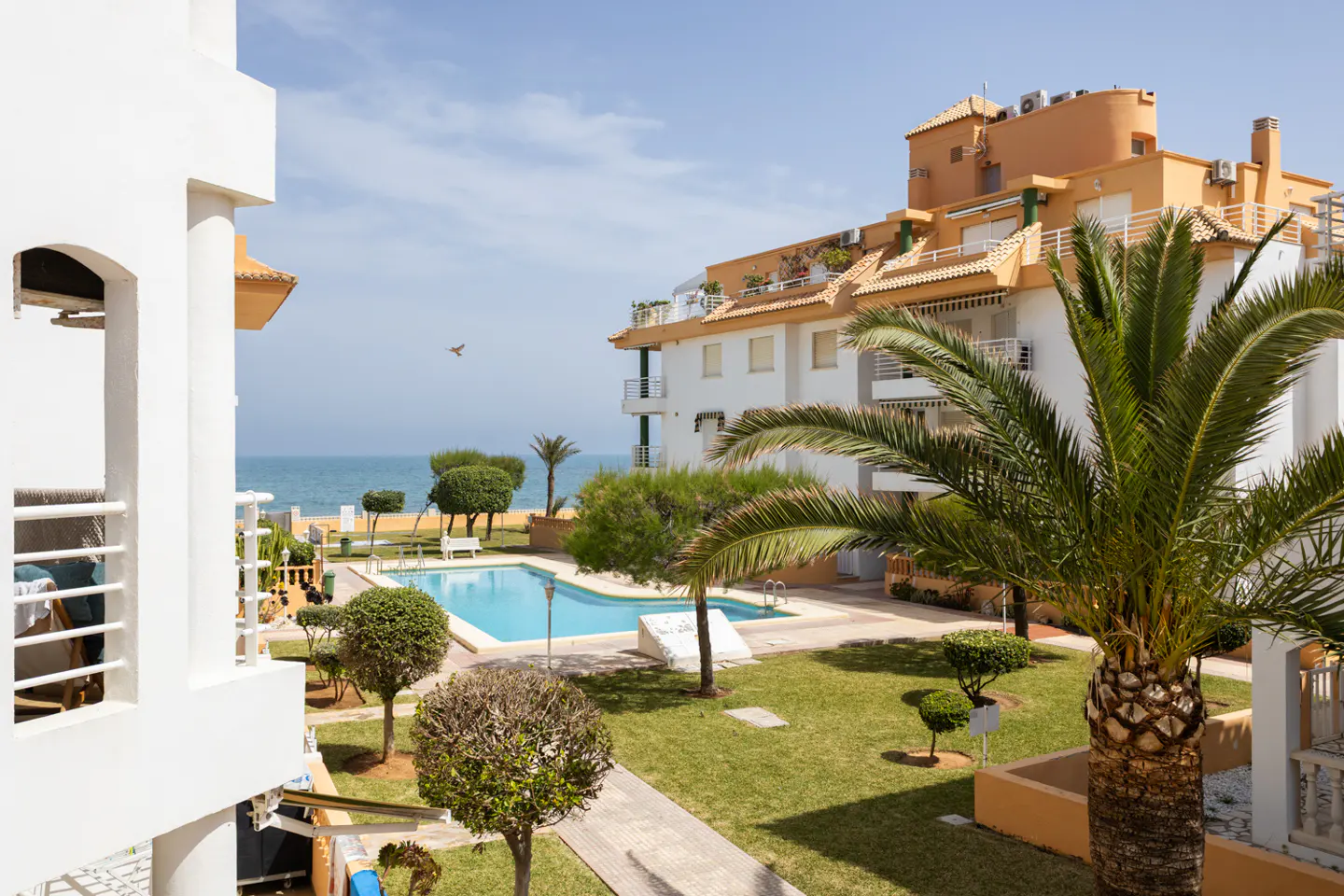 View of a white apartment complex with a pool, lawn, and palm trees near the ocean under a blue sky.