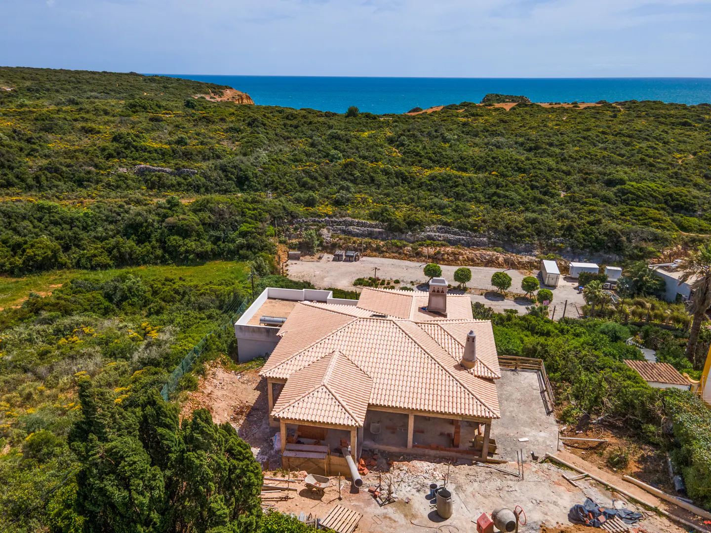 Aerial view of a house under construction with a terracotta tile roof, surrounded by green vegetation and the blue ocean in the background.