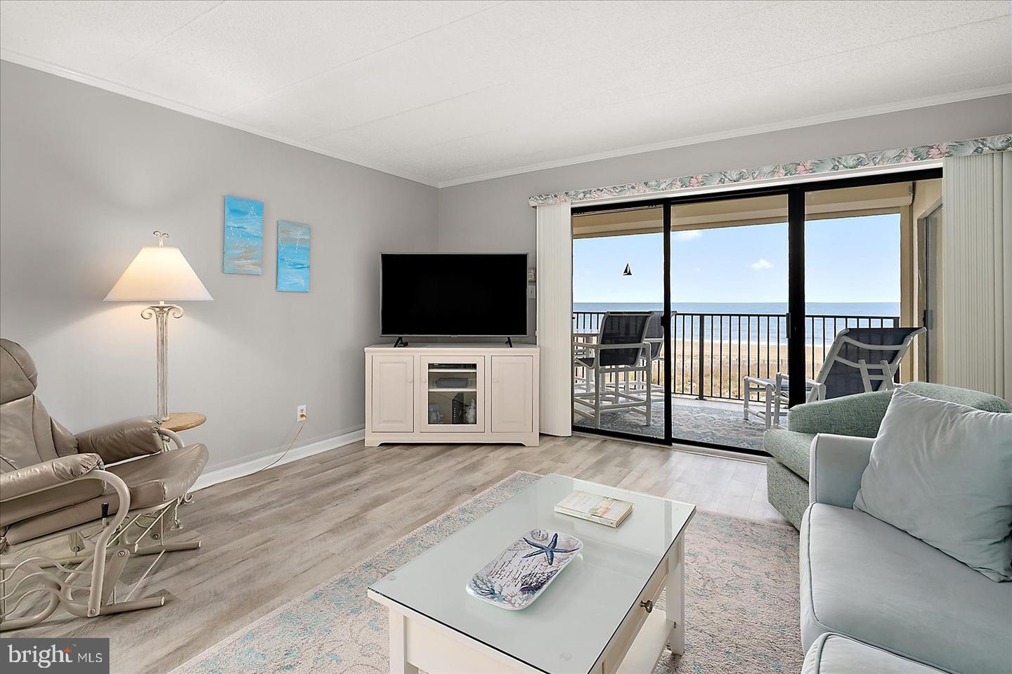 Living room with ocean view. Sliding glass doors open to a balcony with chairs. TV on a white cabinet. Gray walls and wood floors.
