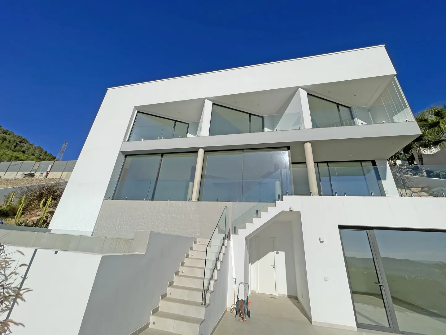 Modern white two-story villa with large glass windows, a glass-railed staircase, and a clear blue sky background.