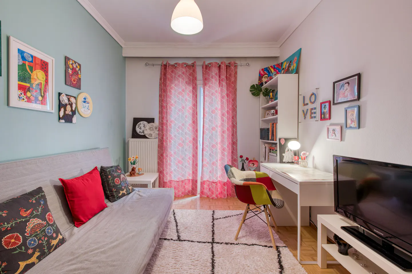 Bright room with a gray sofa, colorful pillows, a white desk, and a TV. Pink curtains hang in front of a window. Artwork adorns the walls.