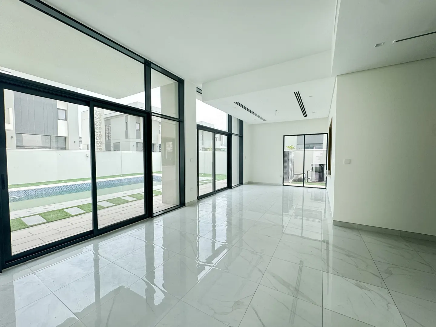 Bright, empty room with white walls and marble floors. Large black-framed windows overlook a pool and backyard.
