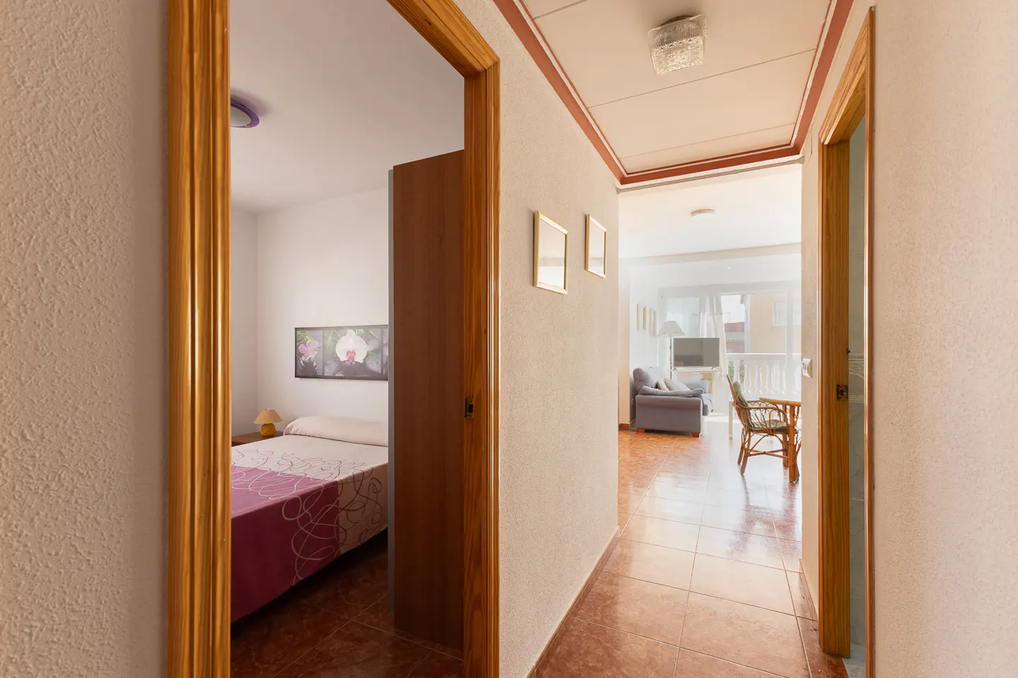 A hallway with tile floors leads to a living room with a blue couch and a bedroom with a bed and a painting.
