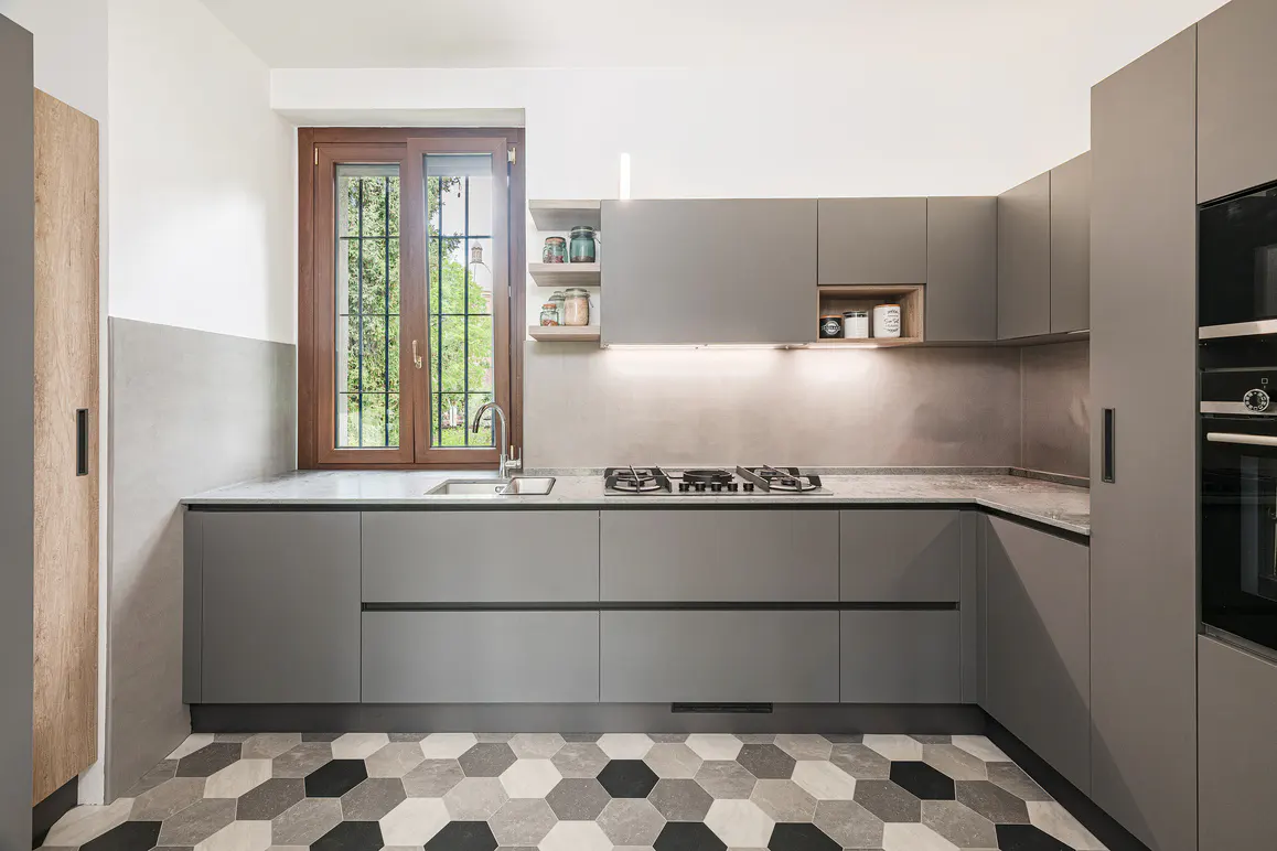 Modern kitchen with gray cabinets, marble countertops, and a geometric tile floor. A window overlooks greenery.