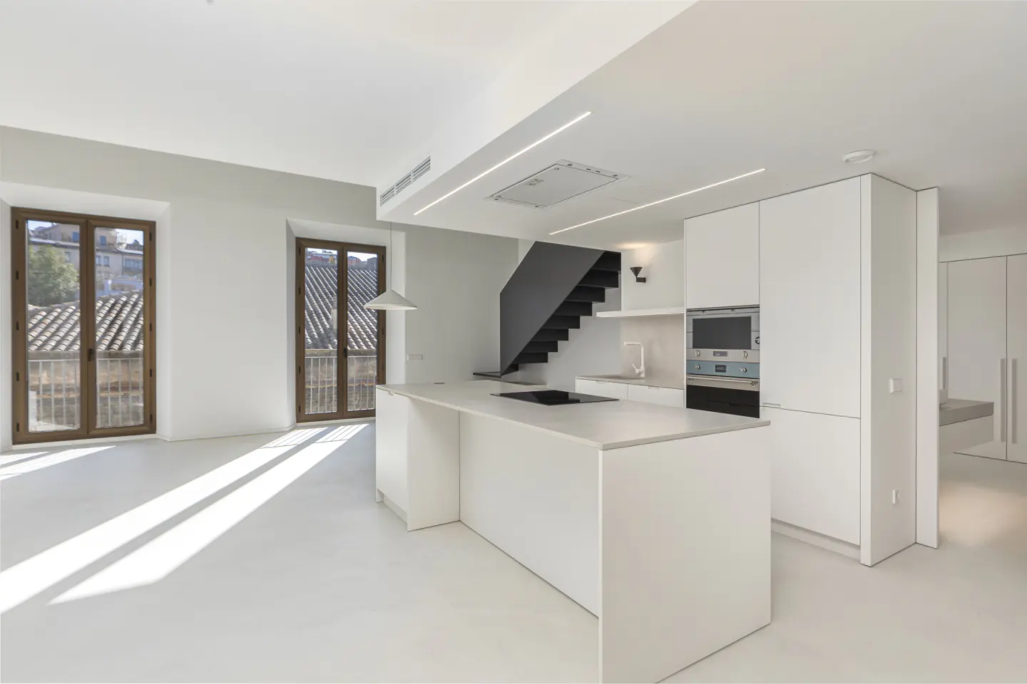 Bright, modern kitchen with white cabinets, island, and floor. Dark stairs lead up. Two windows show a cityscape.