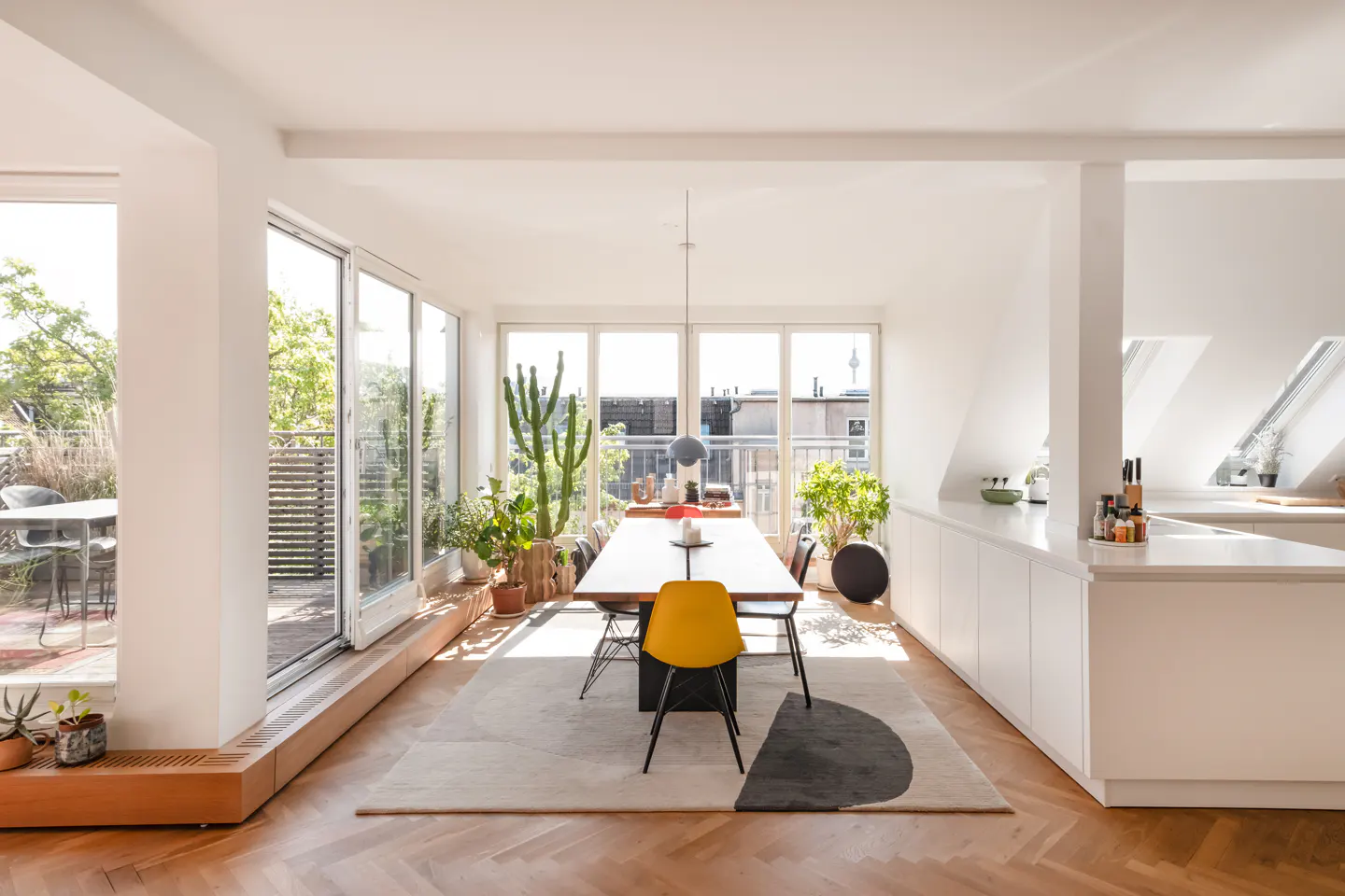 Bright, modern dining area with a long wooden table, black chairs, and a yellow accent chair. Large windows overlook a balcony with plants and city views.