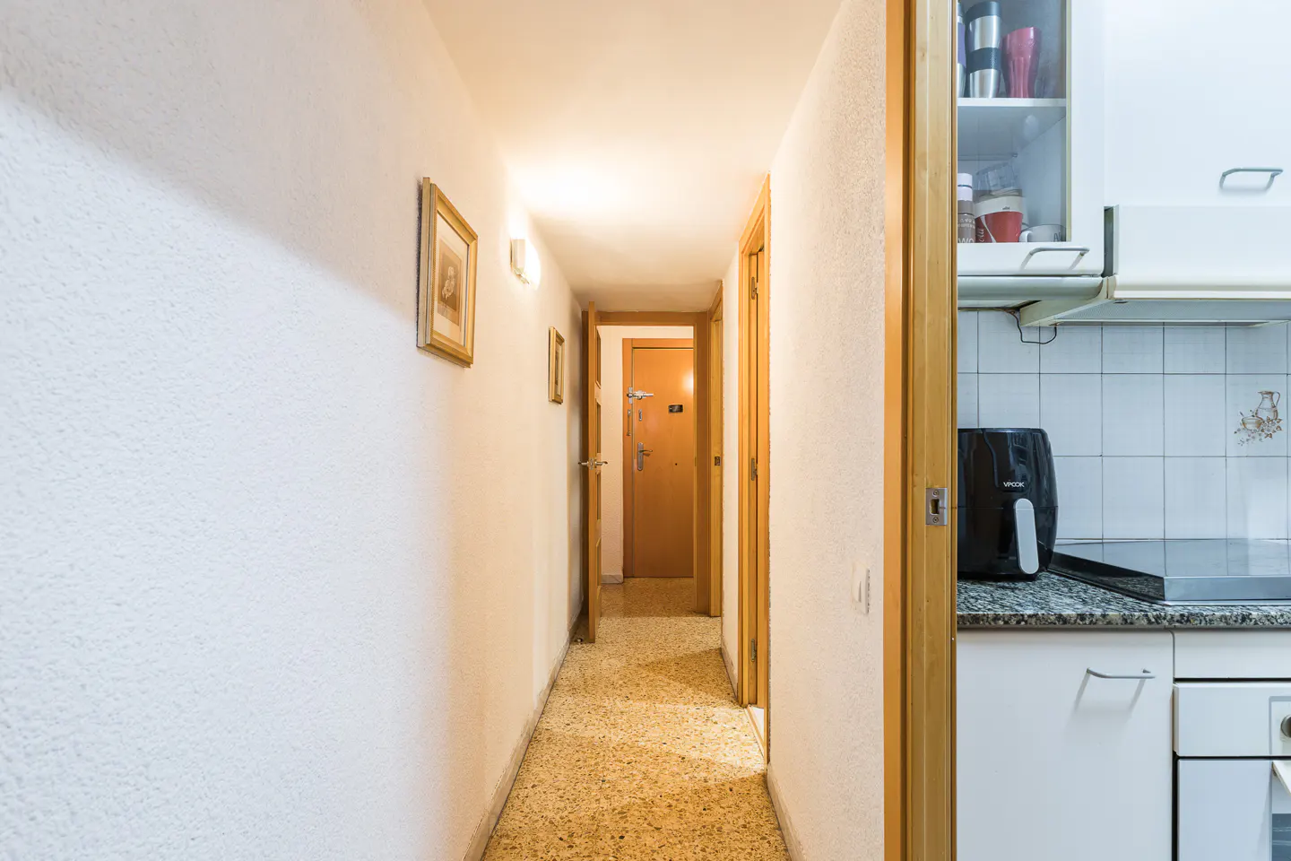 A bright hallway with white walls, a speckled floor, and wooden doors leading to other rooms. A kitchen area is visible on the right.