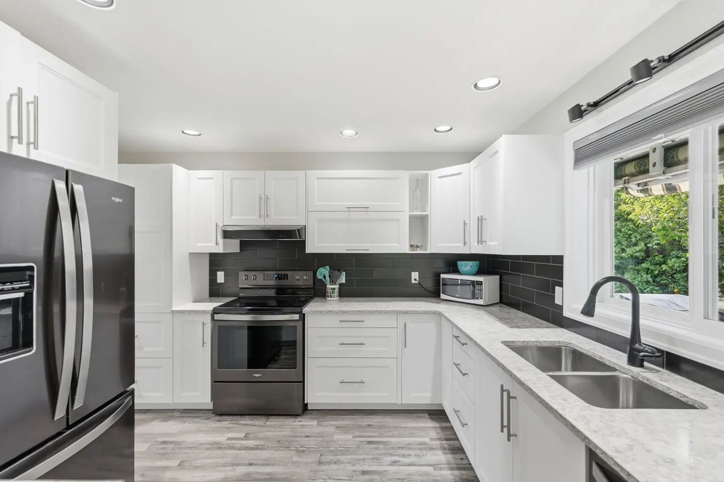 Bright kitchen with white cabinets, gray countertops, and stainless steel appliances. A window overlooks green foliage.