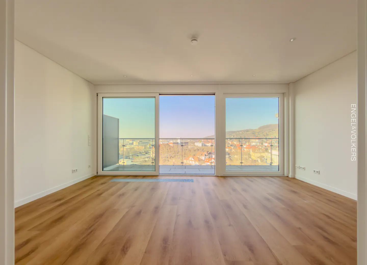 Empty room with wood floors and white walls. Sliding glass doors open to a balcony with a city view.