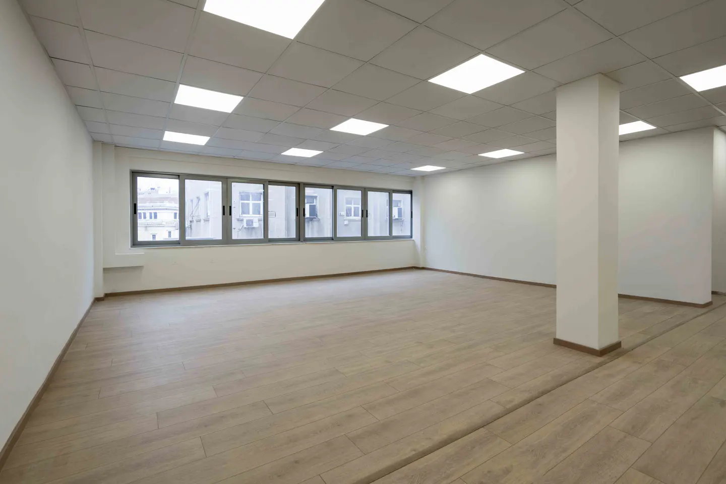 Bright, empty office space with light wood floors, white walls, and a row of windows. Square ceiling lights illuminate the room.