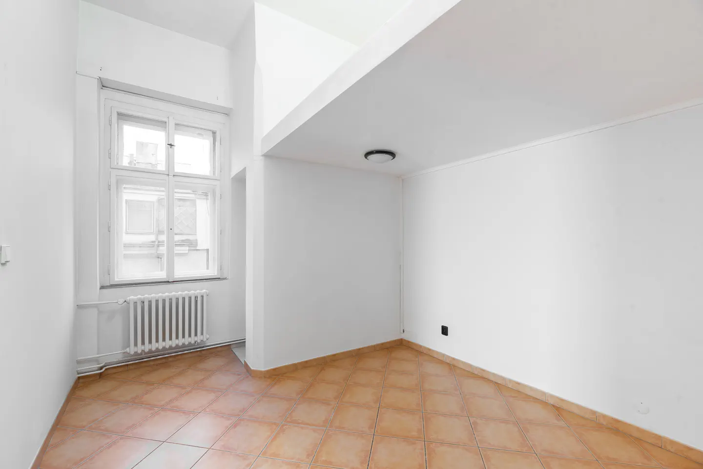 Bright, empty room with white walls, a window with a radiator, and brown tile flooring.