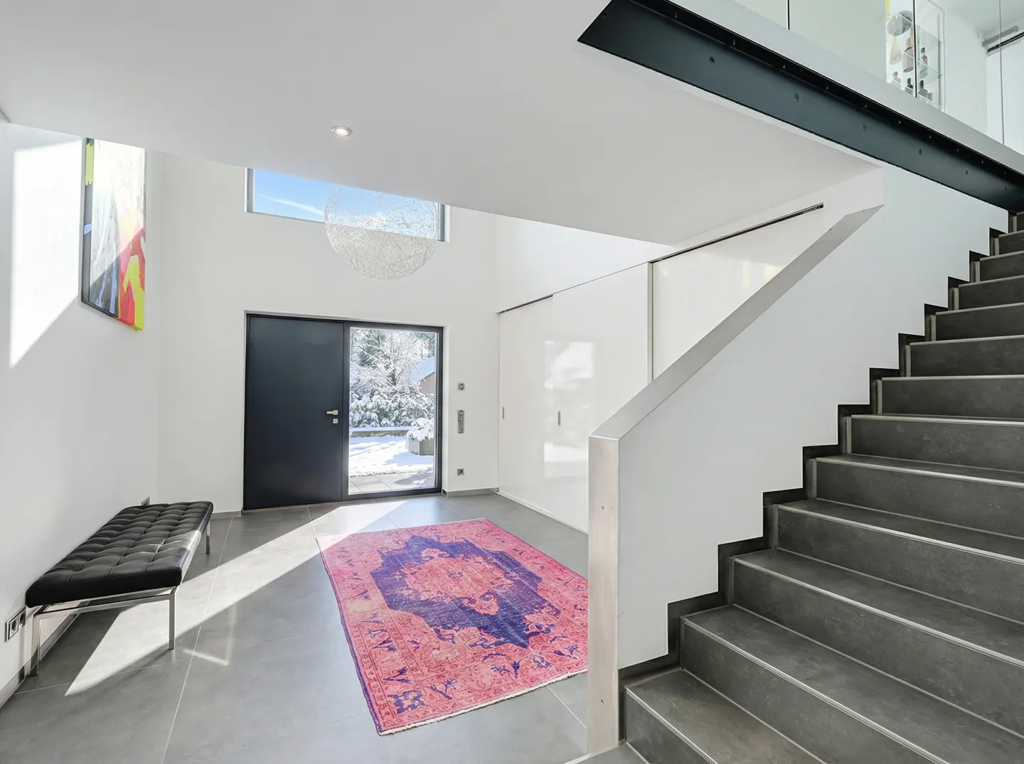 Bright foyer with gray tile floor, black front door, and gray stairs. A pink and blue rug sits in the center. A black bench is on the left.