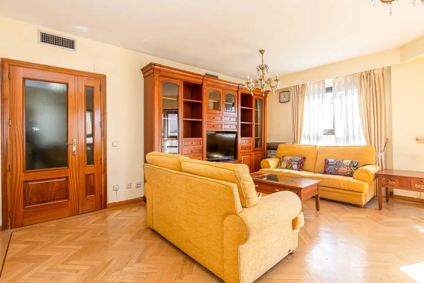Living room with yellow sofas, wood floors, and a large wooden entertainment center. A chandelier hangs from the ceiling.