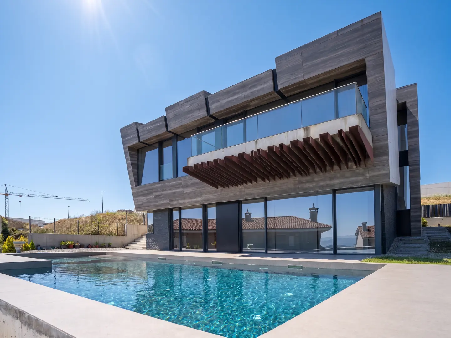 Modern gray house with a pool. Large windows reflect a nearby house. Blue sky.