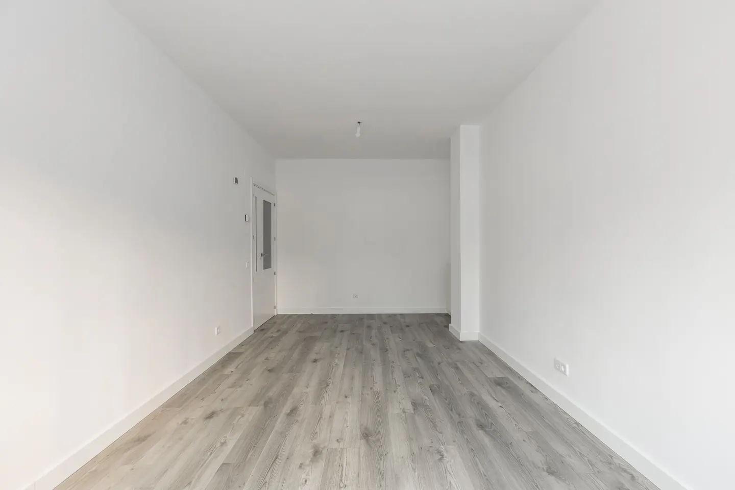Empty room with white walls, gray wood-look flooring, and a white door on the left.