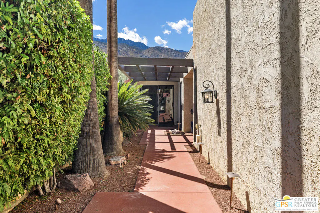 Landscaped walkway to a home's entrance. Palm trees, green bushes, and mountains in the background. The walkway is red.