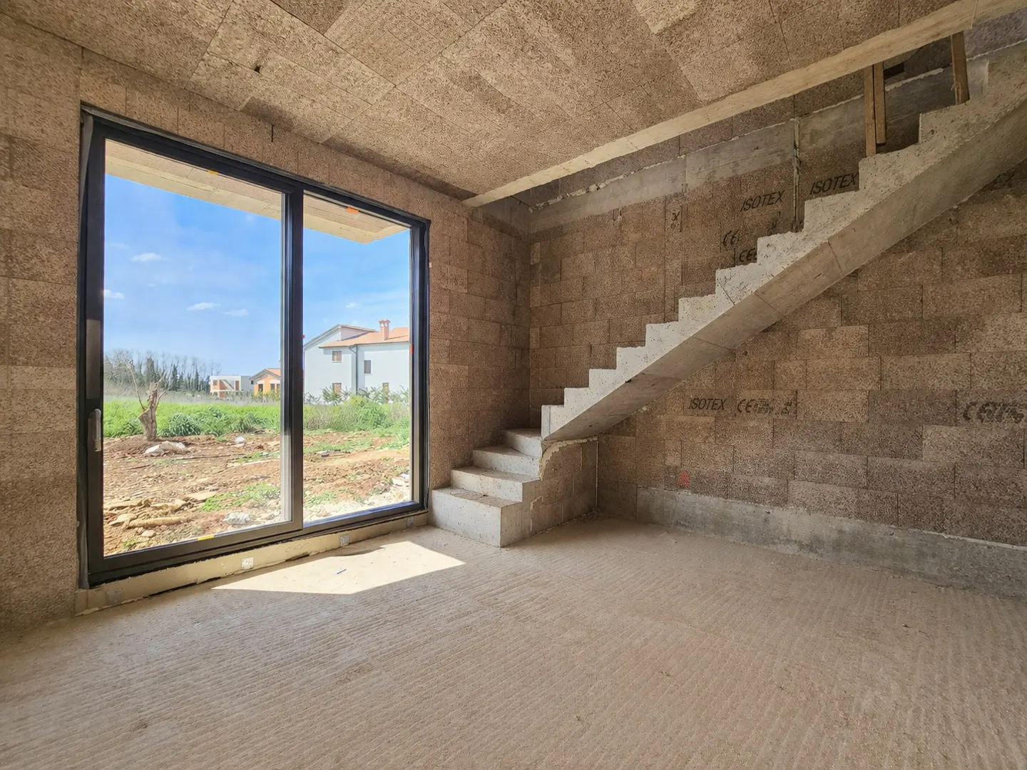 Unfinished room with cork walls, ceiling, and concrete floor. A black-framed sliding glass door shows a yard and house. Concrete stairs lead up.