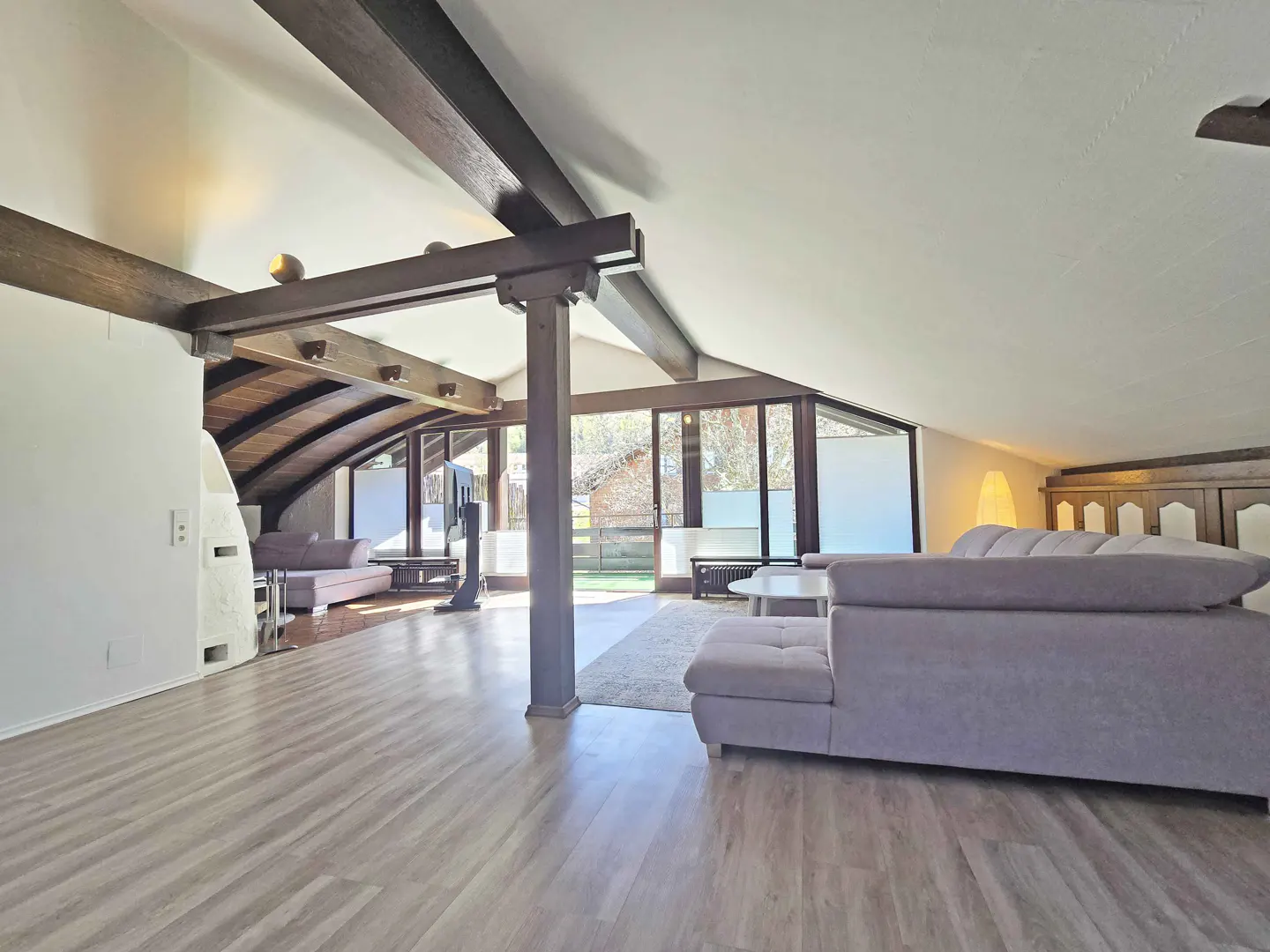 Spacious attic living room with light wood floors, exposed dark wooden beams, and a large grey sectional sofa.