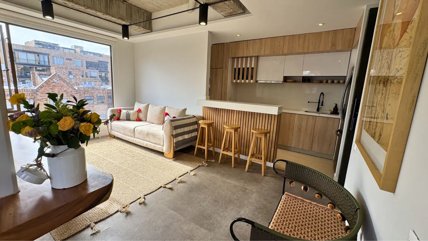 Cozy apartment interior with a beige sofa, wooden stools at a breakfast bar, and a modern kitchen with wood cabinets. A large window offers a city view.
