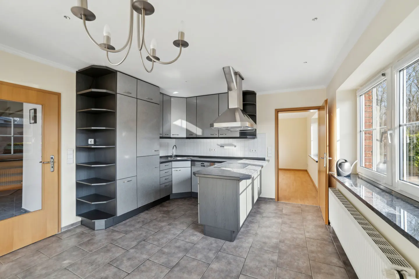 Modern kitchen with grey cabinets, a central island, stainless steel range hood, and tiled floors under a chandelier.