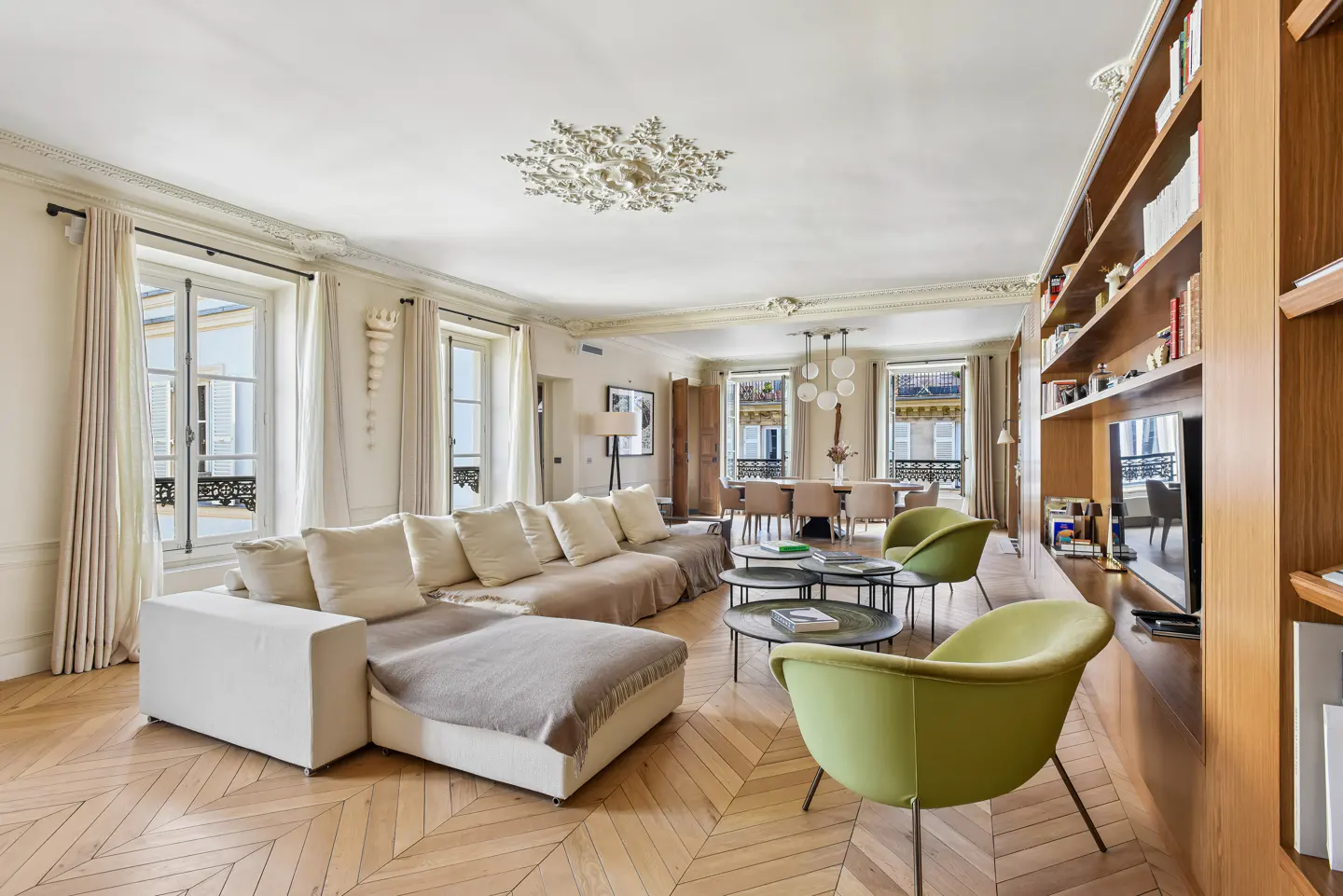Bright living room with herringbone wood floors, white sofa, green chairs, and a dining table visible in the background.