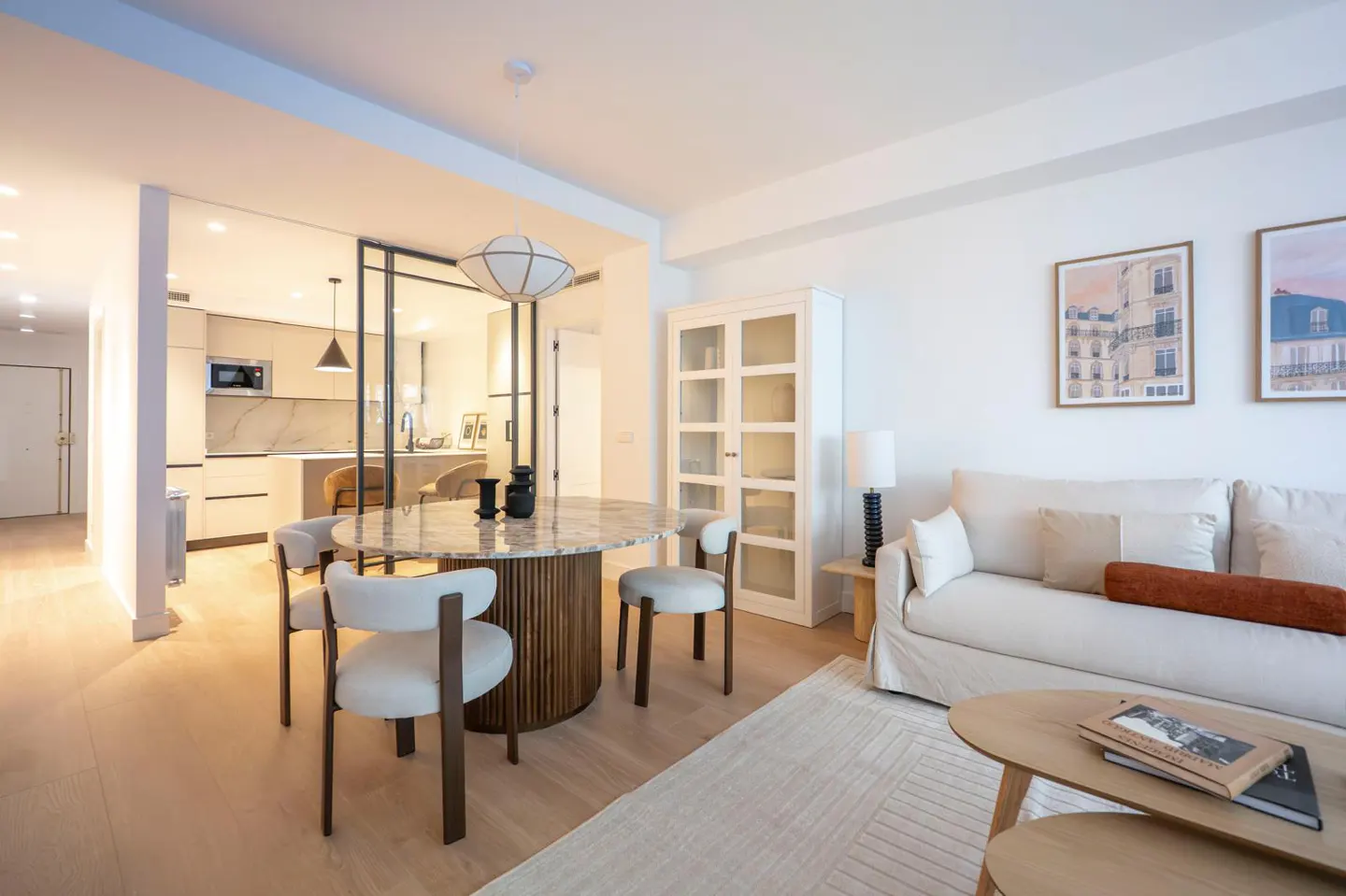 Bright, modern apartment interior. A round marble table with chairs sits near a white sofa, cabinet, and framed art. Kitchen visible in background.
