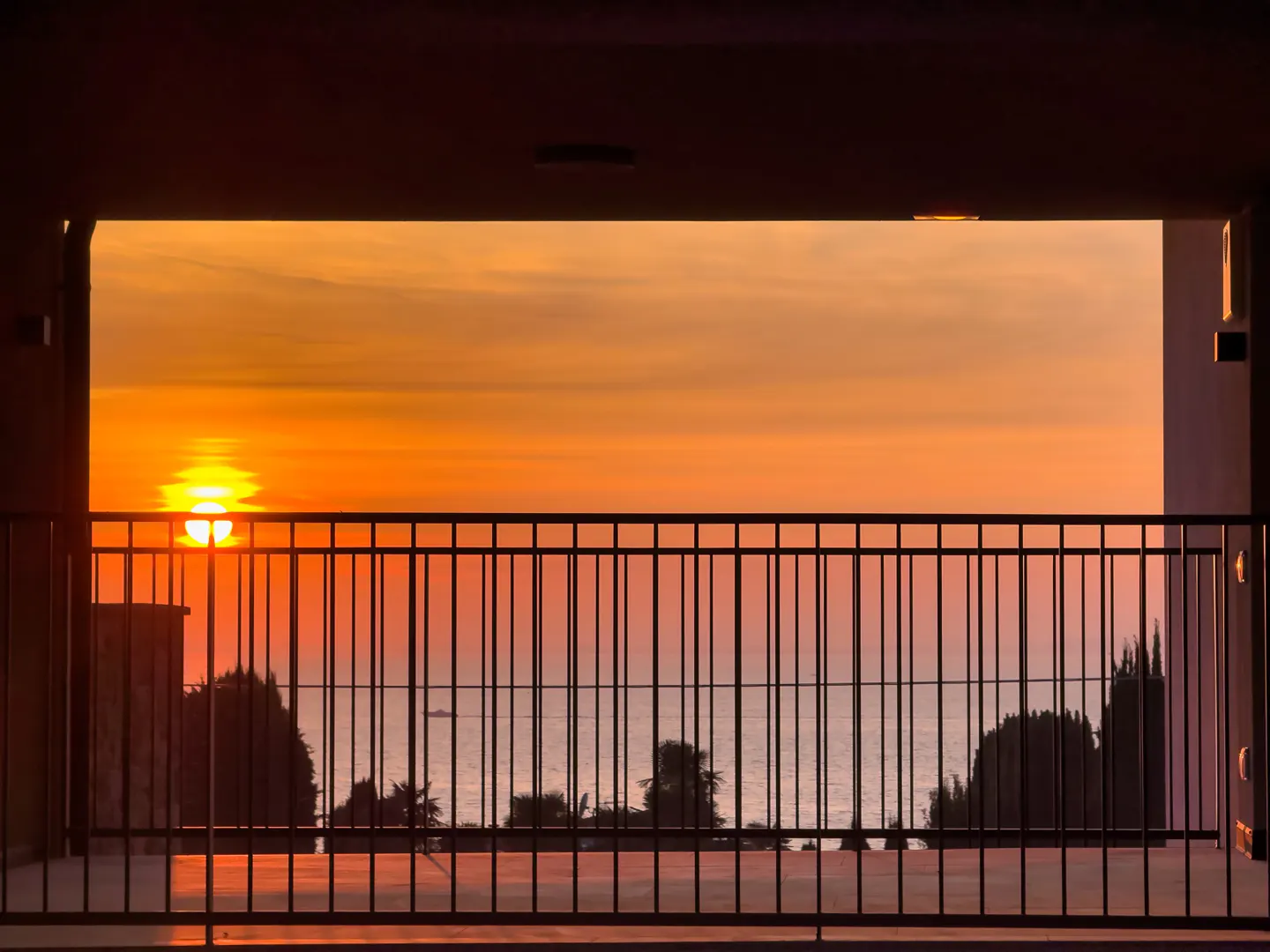 Sunset view from a balcony with a black metal railing, overlooking the sea with trees and a boat. The sky is orange.