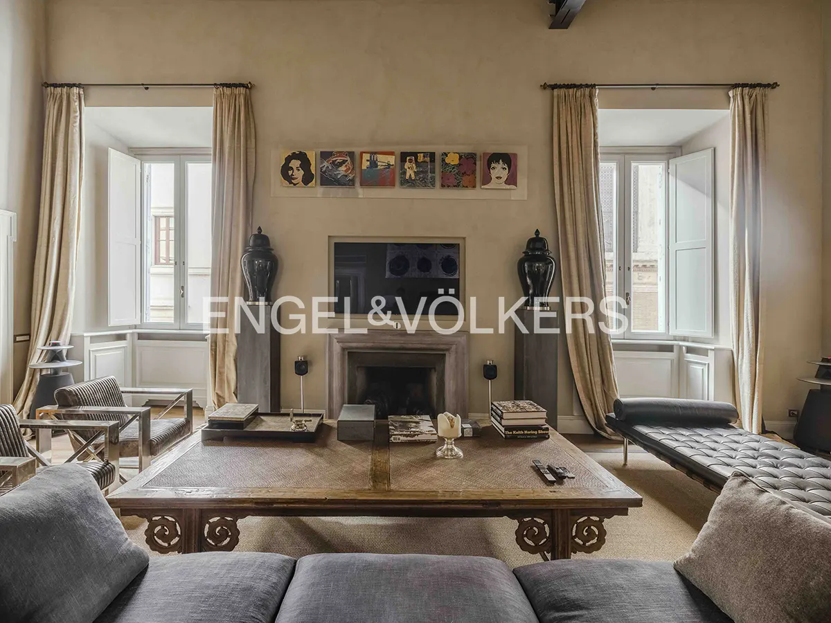 A living room with a large wooden coffee table, fireplace, and two windows with beige curtains. Artwork hangs above the fireplace.