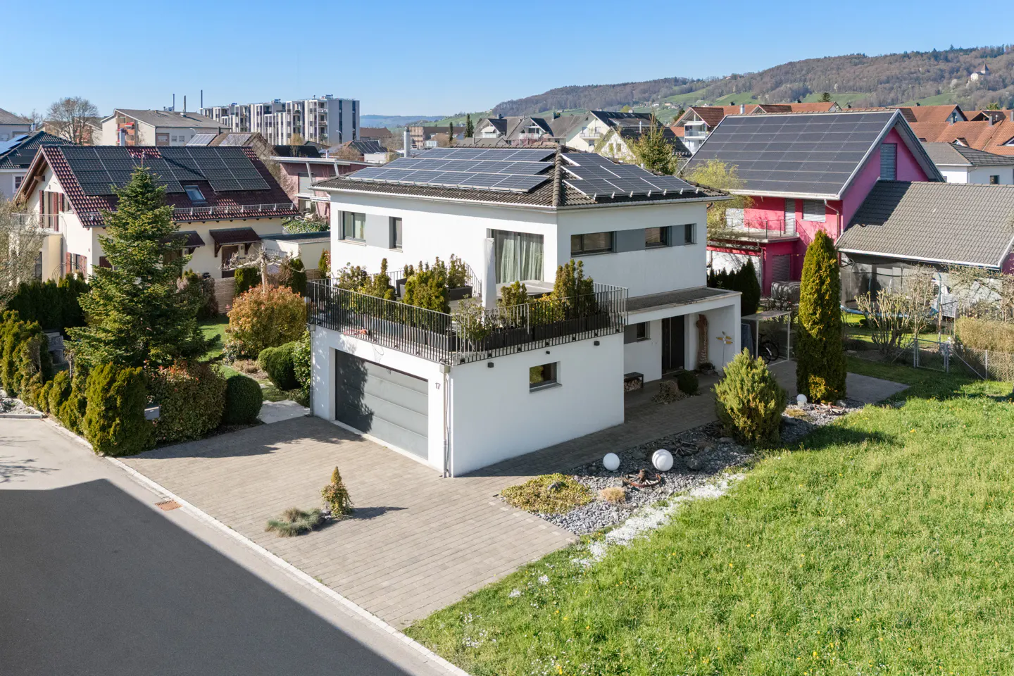 Two-story white house with solar panels, a balcony with plants, and a gray garage door. Green lawn and other houses in the background.