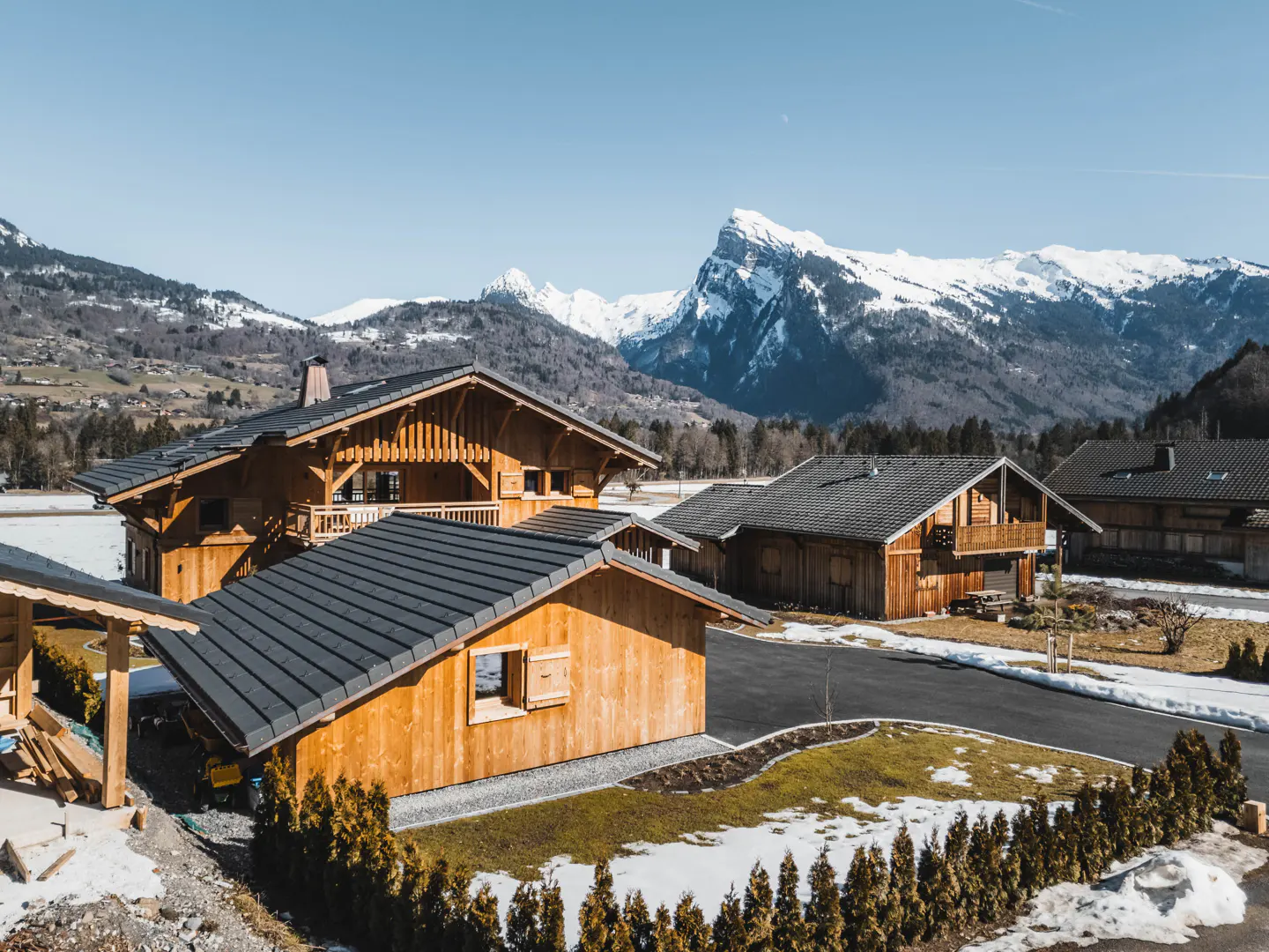 Scenic view of wooden chalets with dark roofs, set against snow-capped mountains under a clear blue sky.