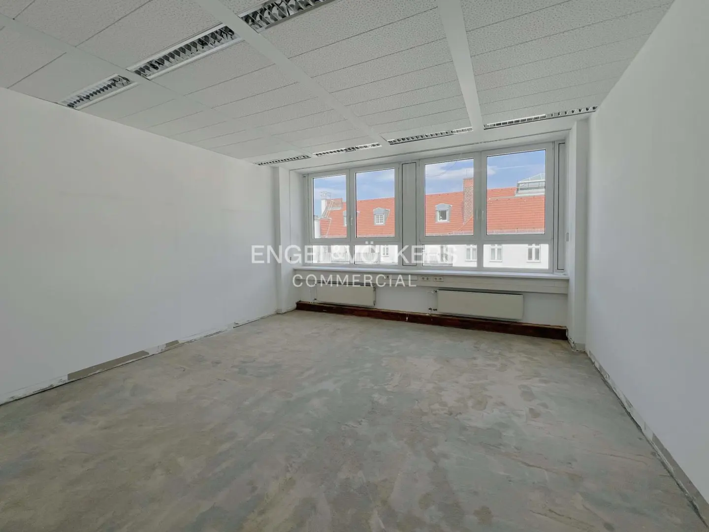 Empty office space with white walls, concrete floor, and large windows overlooking a red-roofed building.