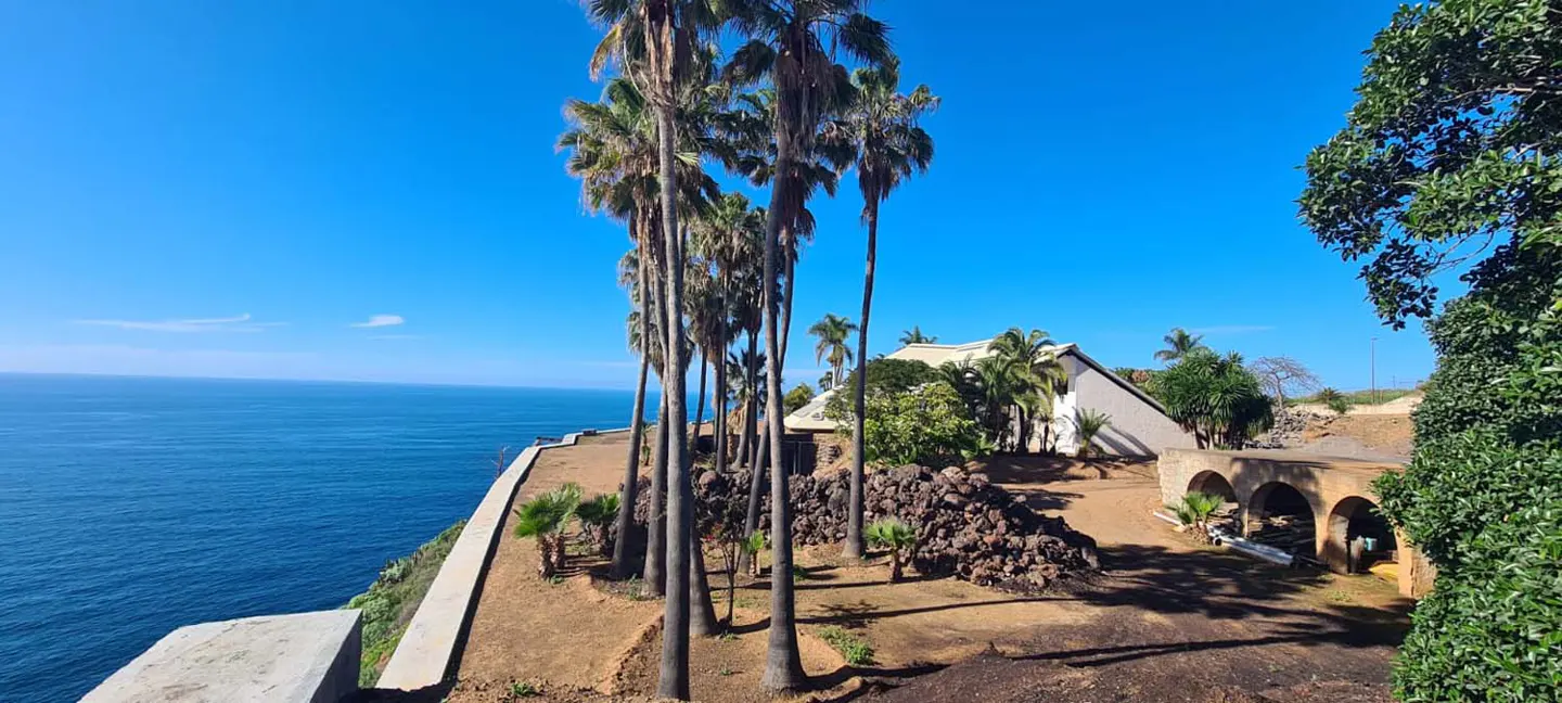 Ocean view property with palm trees. White house with a stone wall and arched structure. Blue sky.