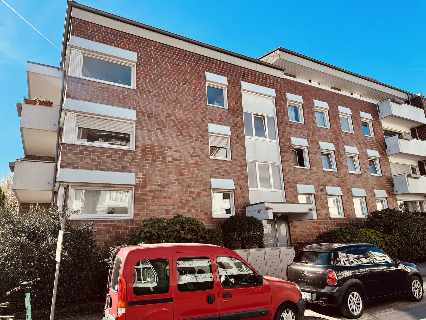 A three-story brick apartment building with white trim, balconies, and two parked cars under a blue sky.
