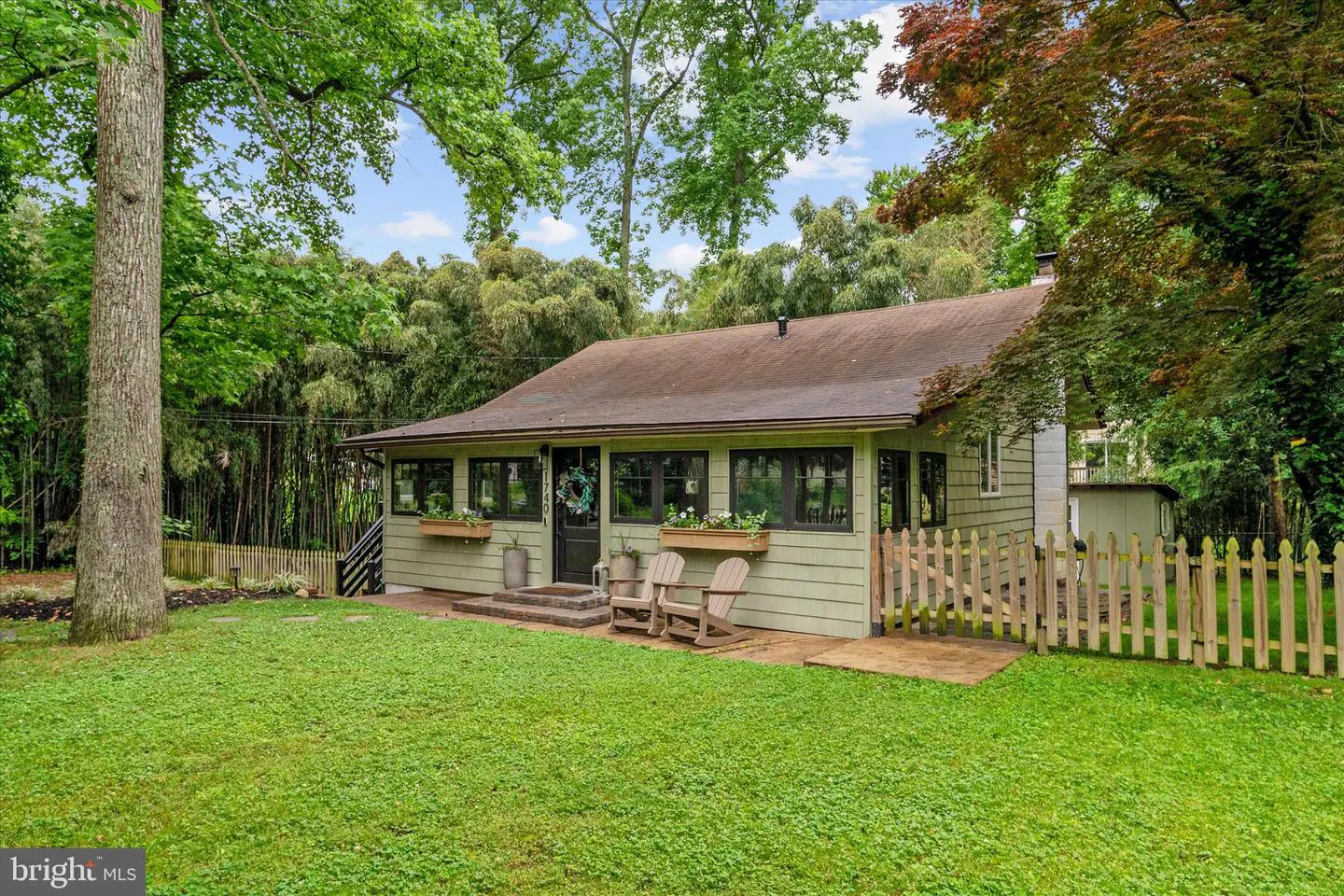Exterior view of a one-story, light green house with a brown roof, surrounded by lush green trees and a lawn. Two wooden chairs sit on the porch.