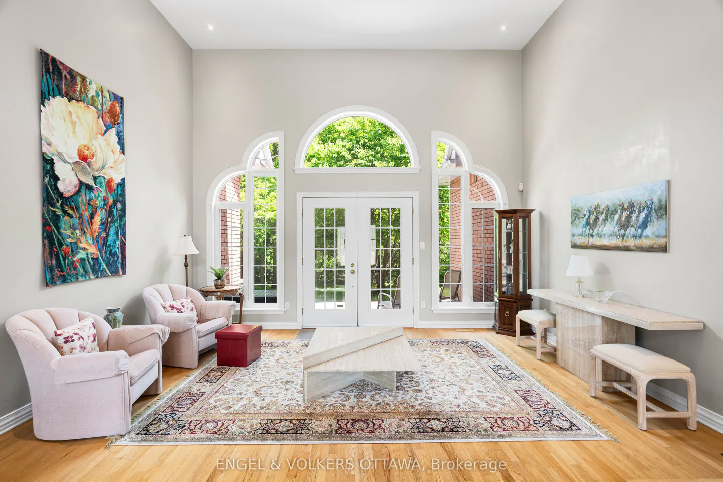 Bright living room with high ceilings, two pink armchairs, a patterned rug, and large arched windows with garden views.