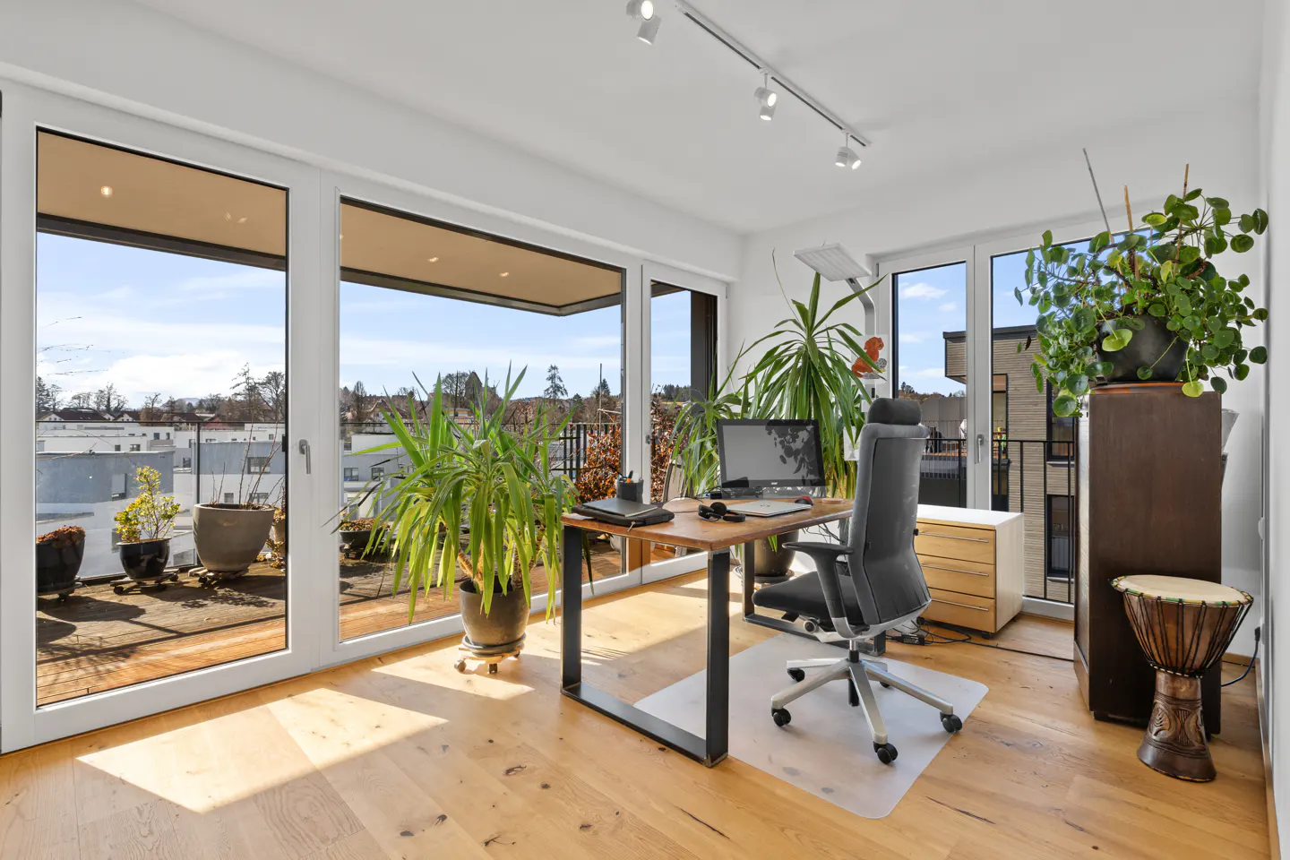 Bright home office with wood floors, a desk, computer, and plants. Large windows open to a balcony with city views.