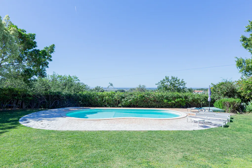 Outdoor pool with lounge chairs on a sunny day. Green grass and trees surround the pool area. Blue sky in the background.