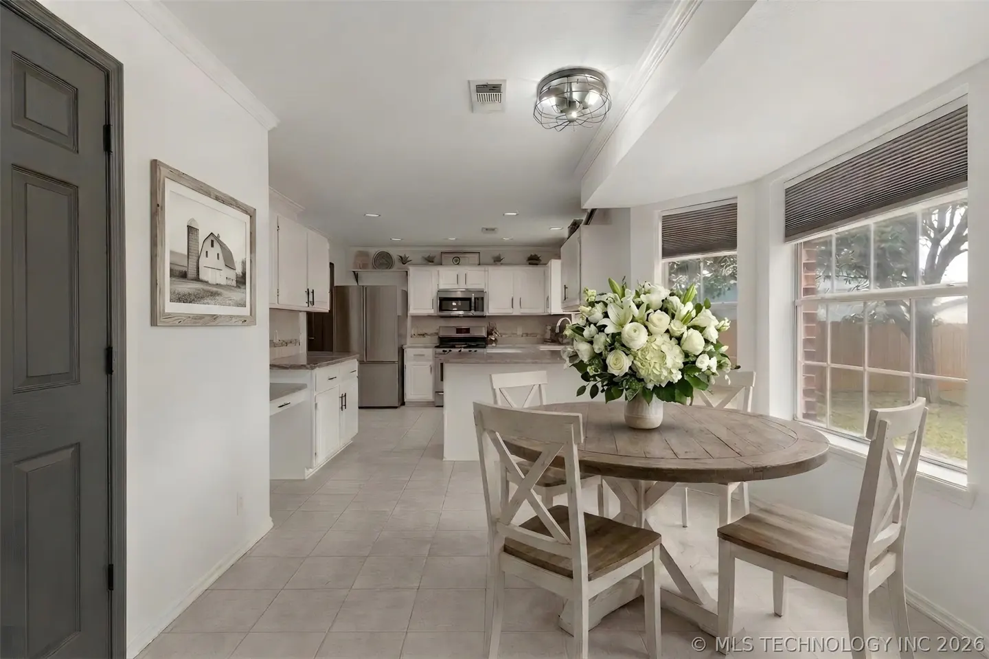 A bright kitchen with white cabinets, stainless steel appliances, and a round wooden table with white chairs. A large bouquet of white flowers sits on the table.