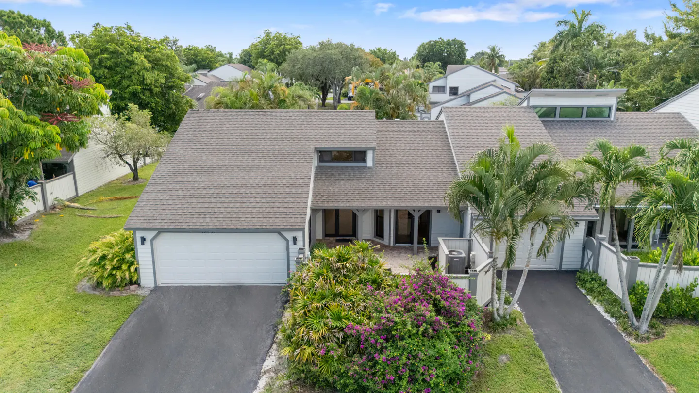 Aerial view of a light blue, two-story home with a gray roof, white garage door, and lush green landscaping.