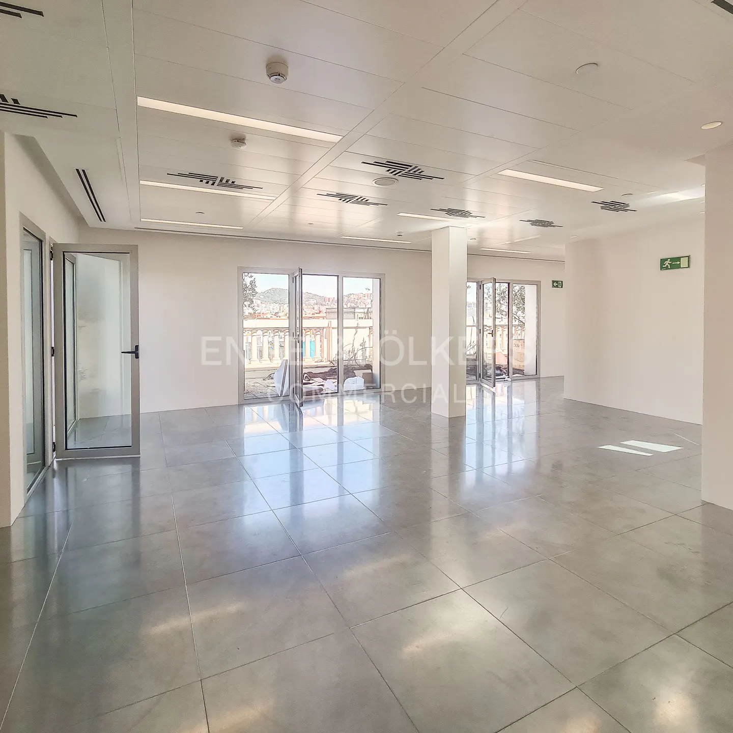 Bright, empty commercial space with gray tile floor, white walls, and glass doors leading to a balcony with city views.