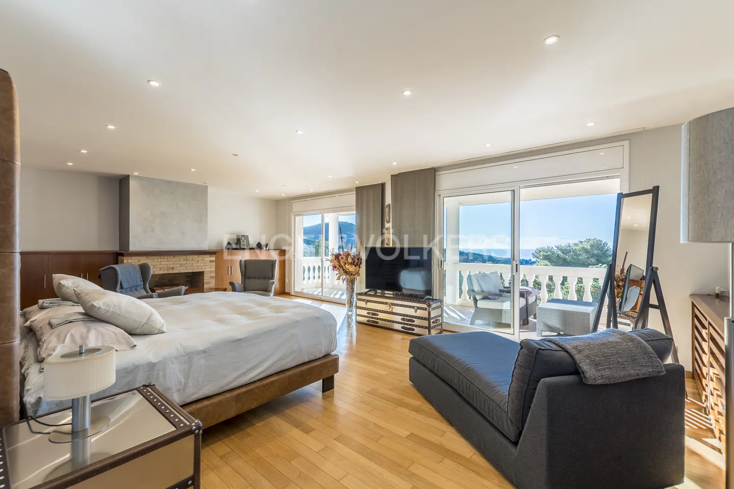 A bright, spacious bedroom with a king bed, fireplace, and balcony overlooking a scenic view. A chaise lounge sits near the balcony doors.