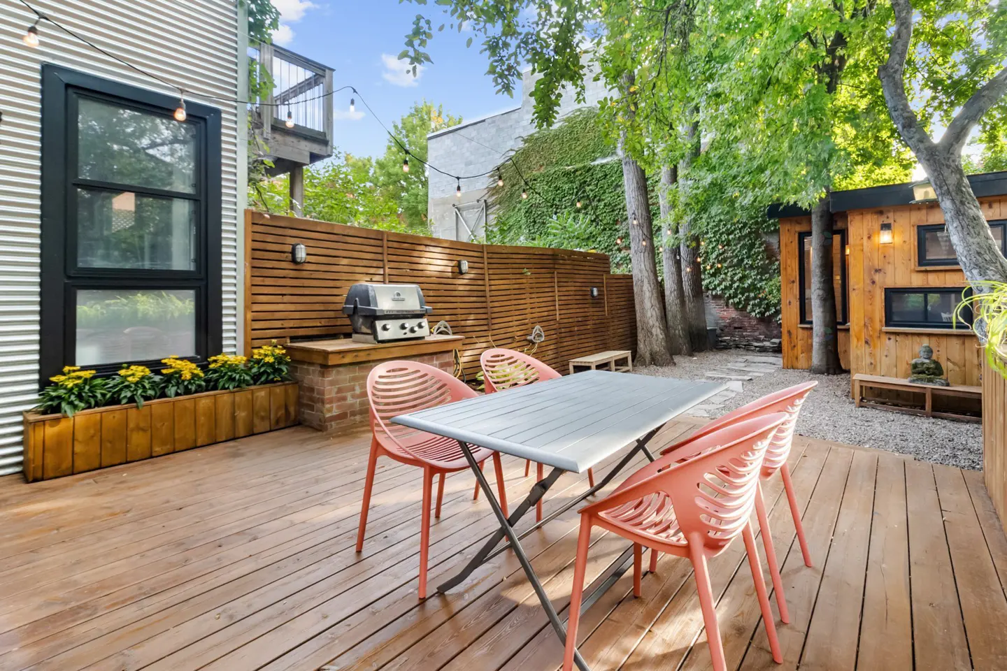 Outdoor patio with a wooden deck, table, pink chairs, grill, and a modern corrugated metal building. String lights hang overhead.