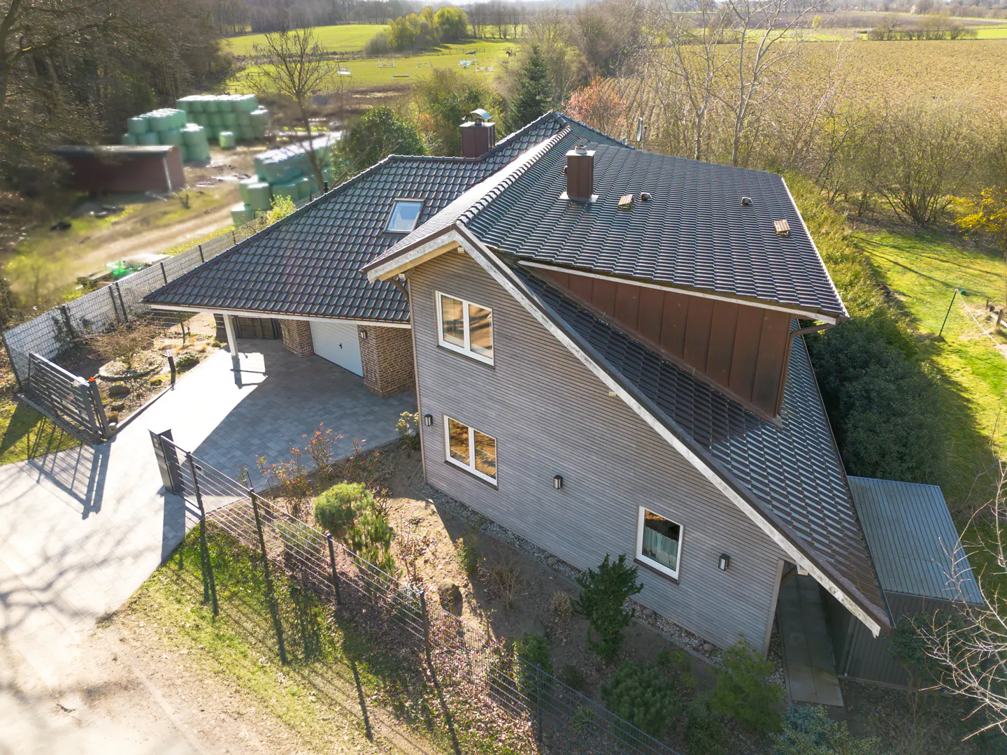 Aerial view of a gray, modern house with a dark roof, surrounded by green grass and trees. A fence runs along the property line.